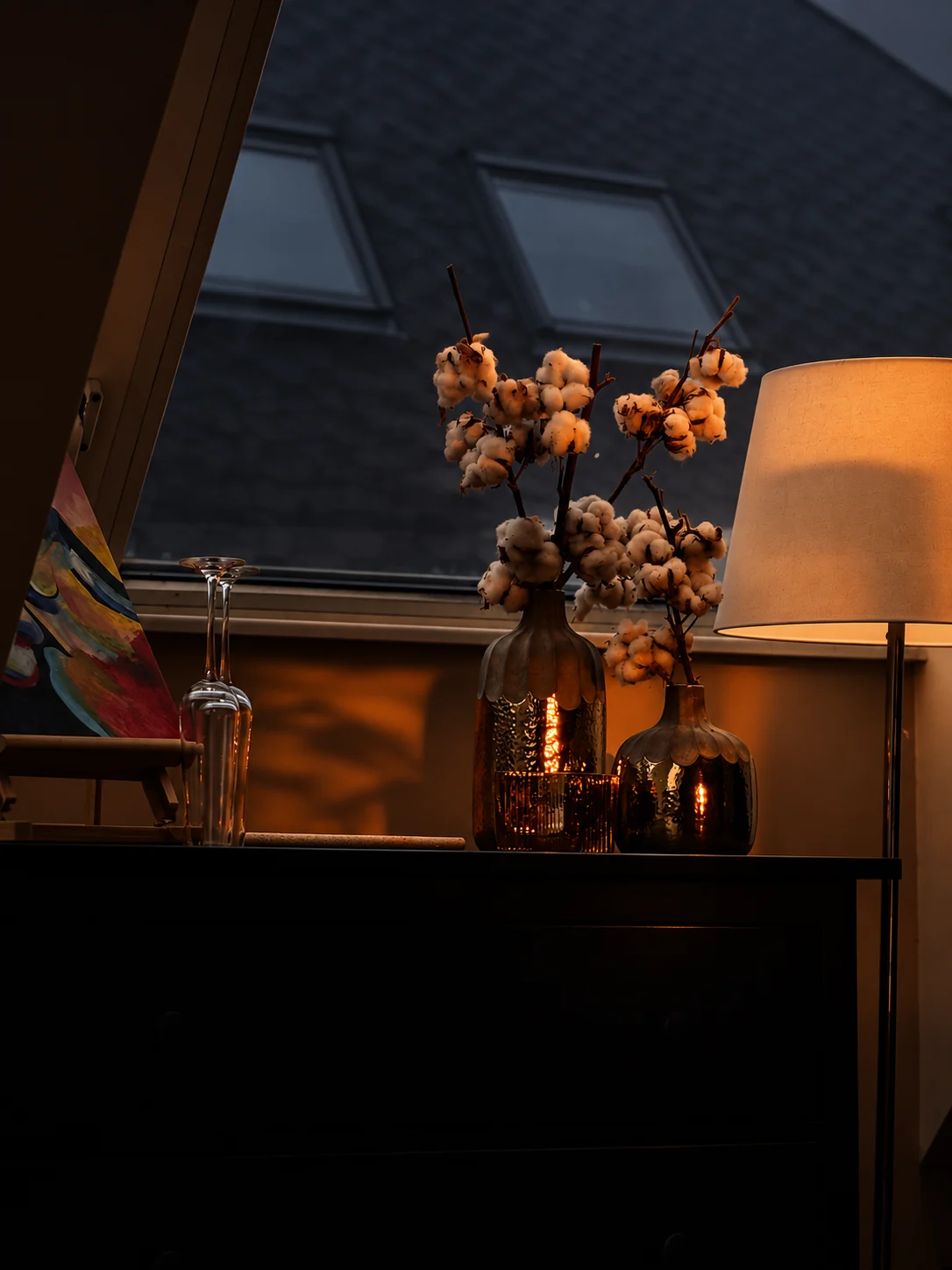 Cotton branches in glass vases by a window at dusk, warm lamplight behind